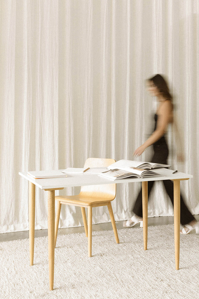 Frau im Hintergrund, ästhetisch in Schwarz gekleidet, geht an Schreibtisch mit Büchern vorbei, minimalistische Atmosphäre